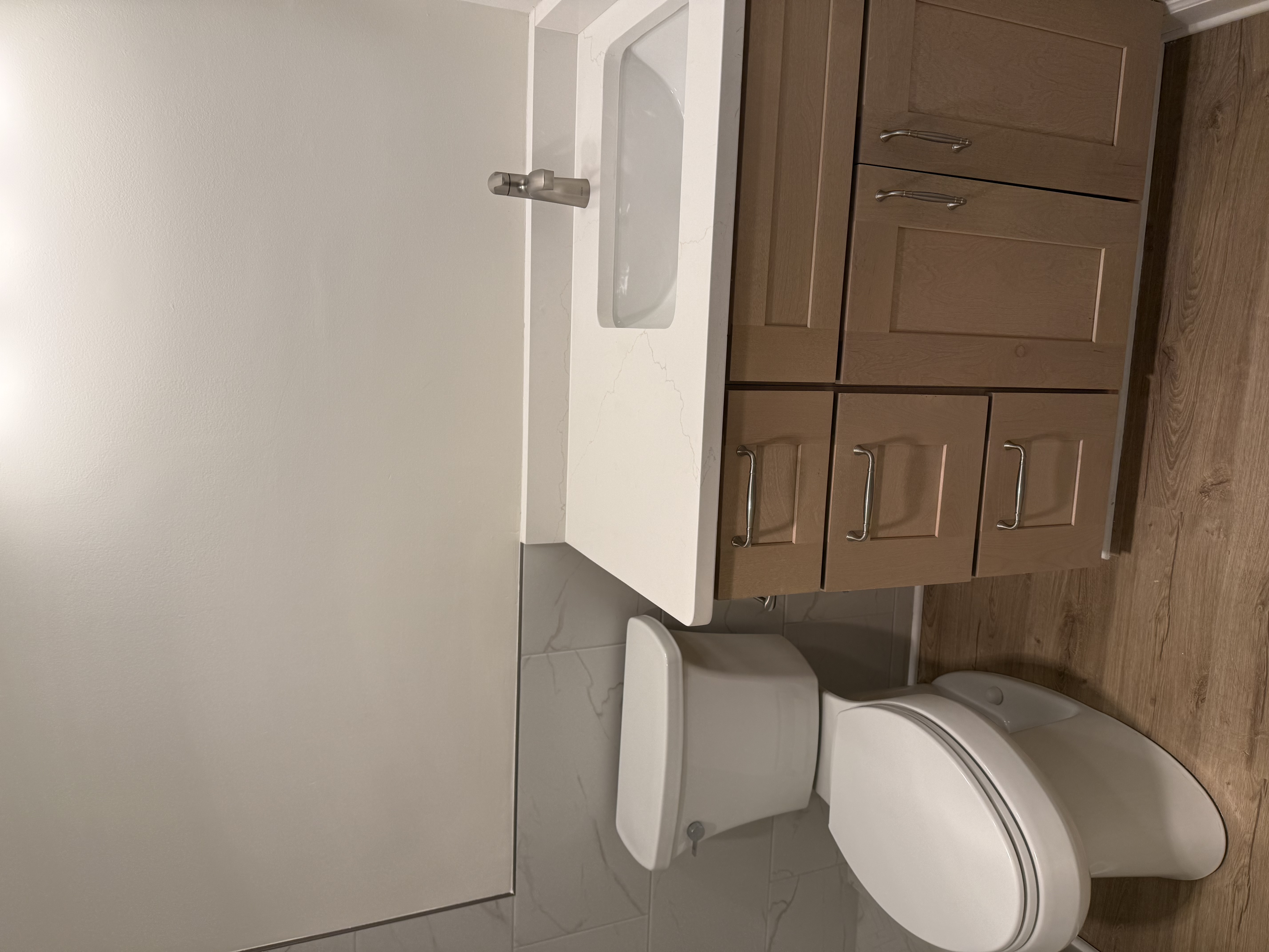 Finishing detail — the new wood-tone vanity with white quartz countertop beside the toilet, set against the gray marble-look tile wainscot and wood-look floor