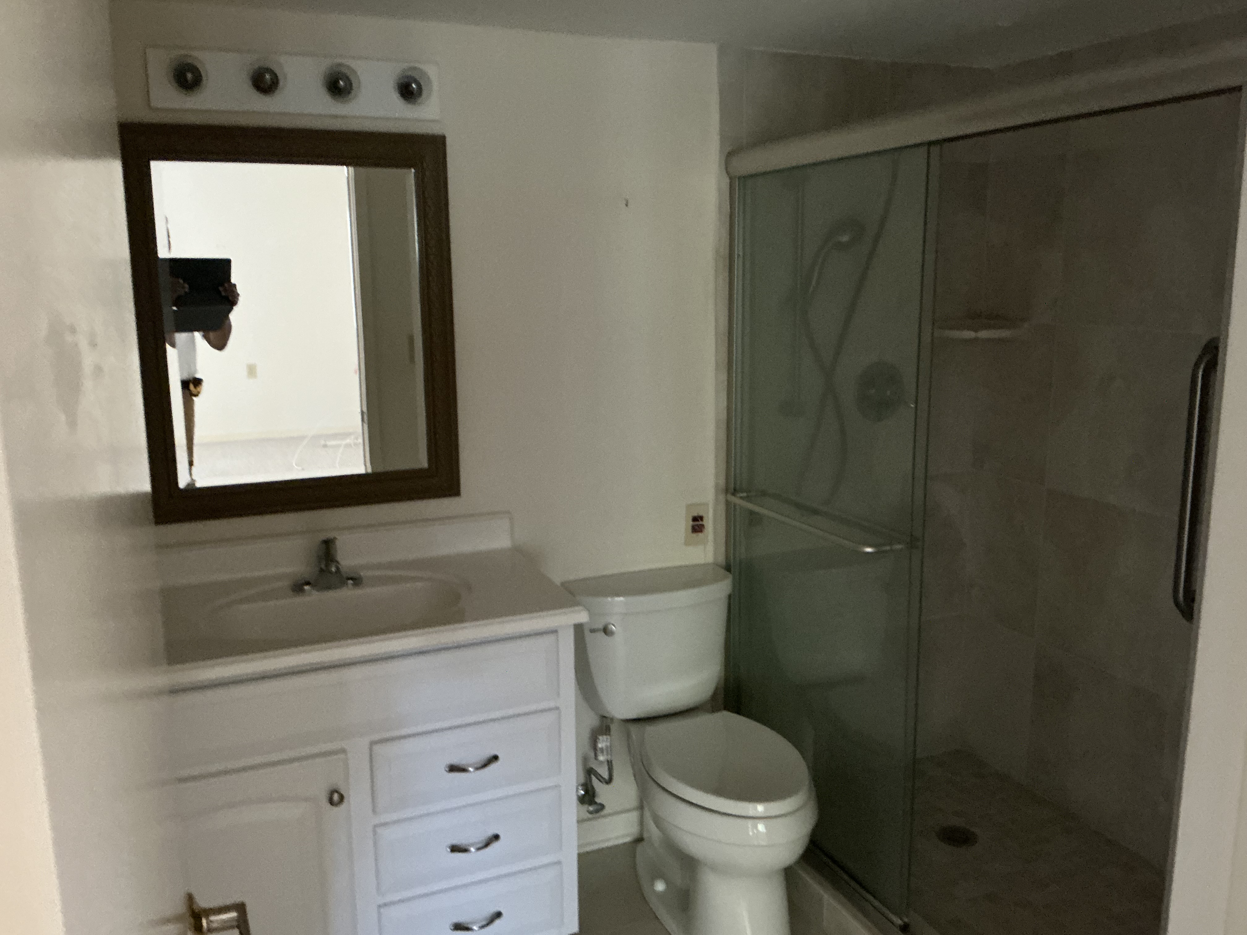 The bathroom as the room began to take shape — white vanity with wood-framed mirror and four-bulb light, toilet and glass-enclosed shower