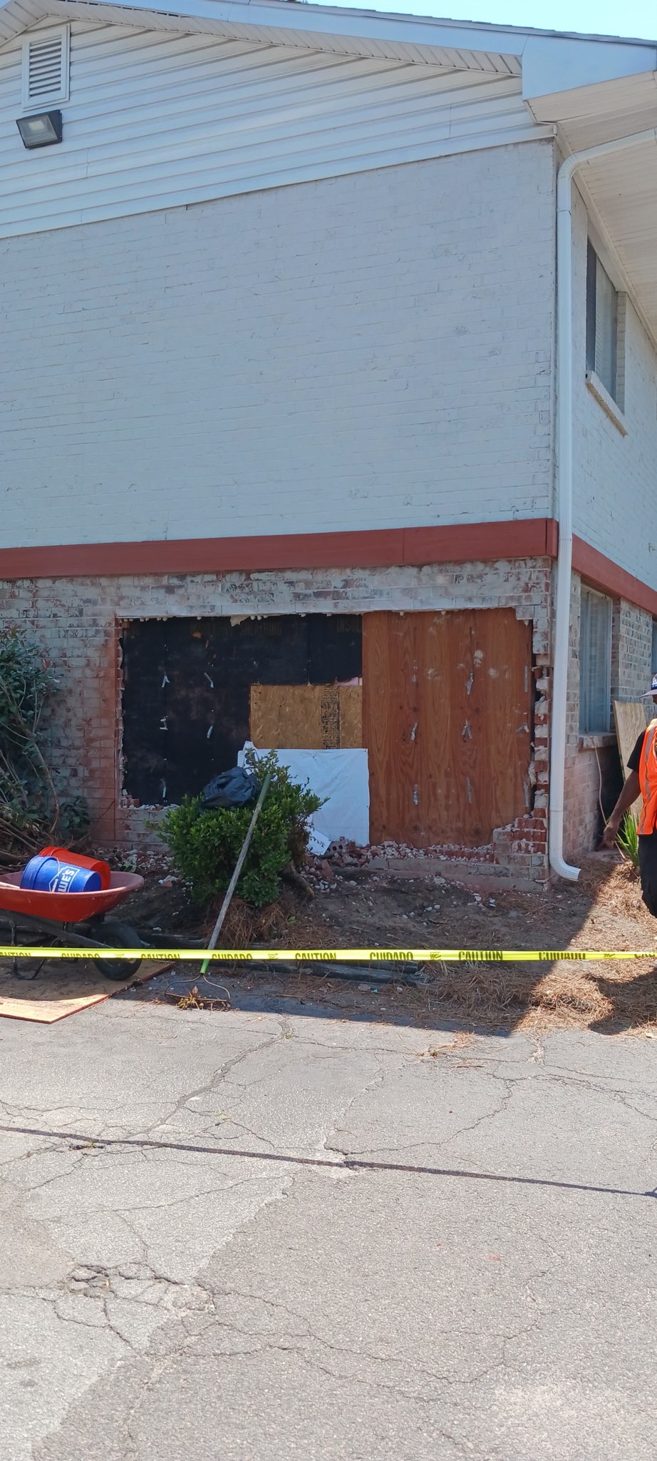 Before the repair — the opening boarded and covered at ground level with plywood, black weather material and cardboard, caution tape across the front, a wheelbarrow against the wall and a crew member in a safety vest working at the corner