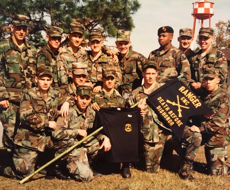 Fourteen soldiers in U.S. Army woodland camouflage in three rows outdoors with the folded Ranger guidon reading Death Before Dishonor, dry grass and evergreens behind them