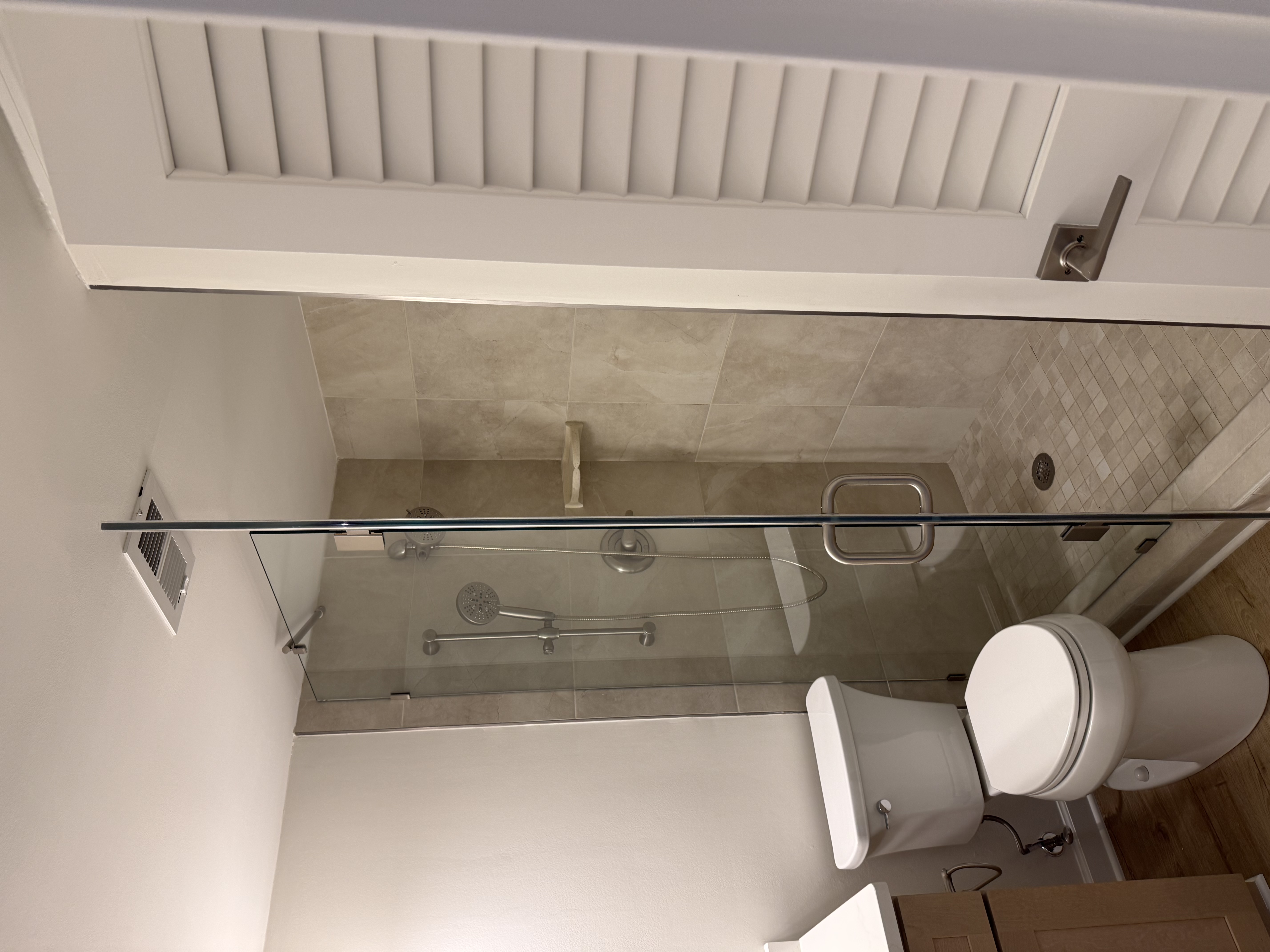 Finished walk-in shower seen from the front — frameless glass enclosure, tan tile walls and mosaic shower floor, with the toilet in the foreground