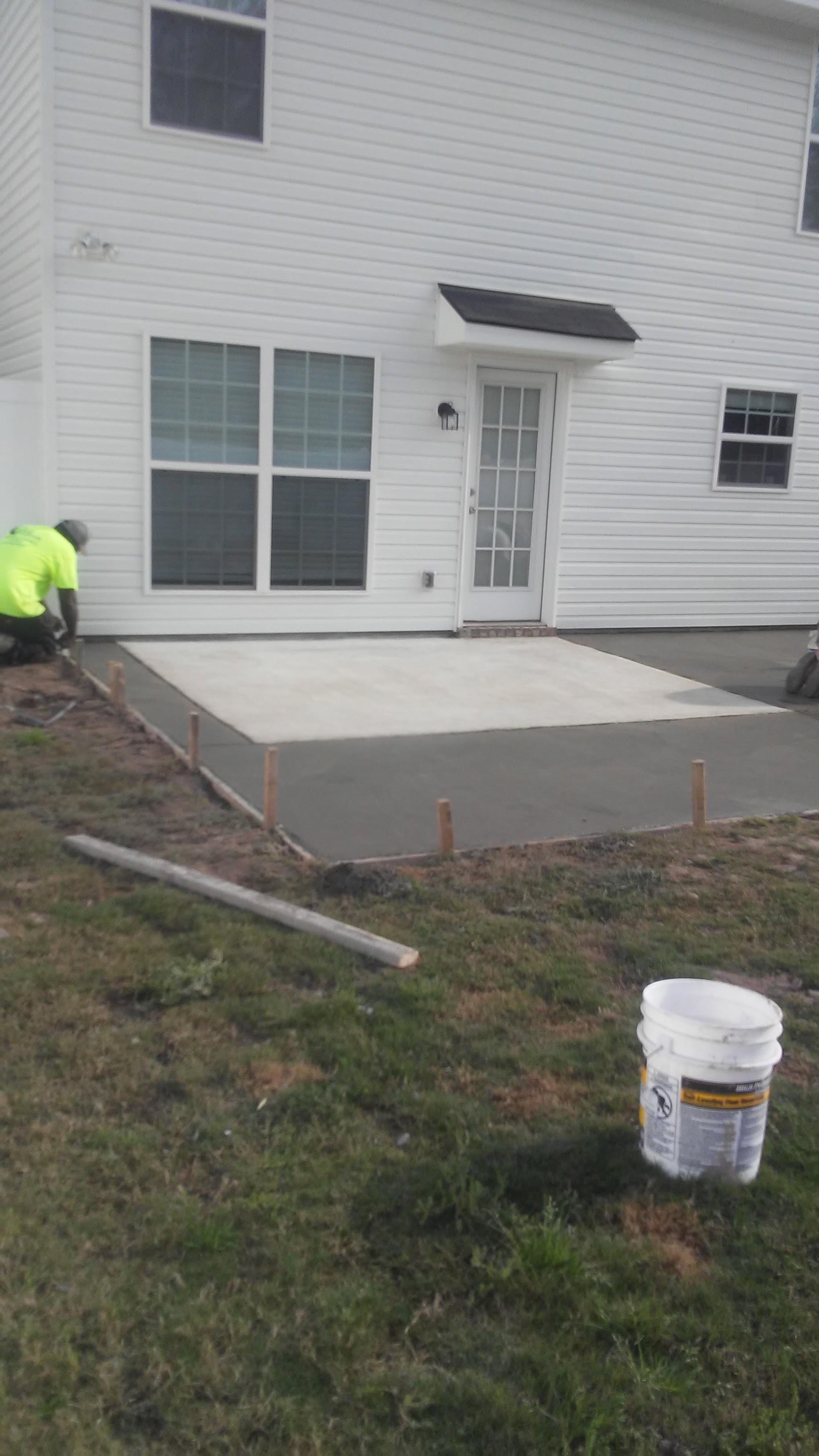 The new patio slab curing behind the grade stakes, against the house’s grid window and glass door