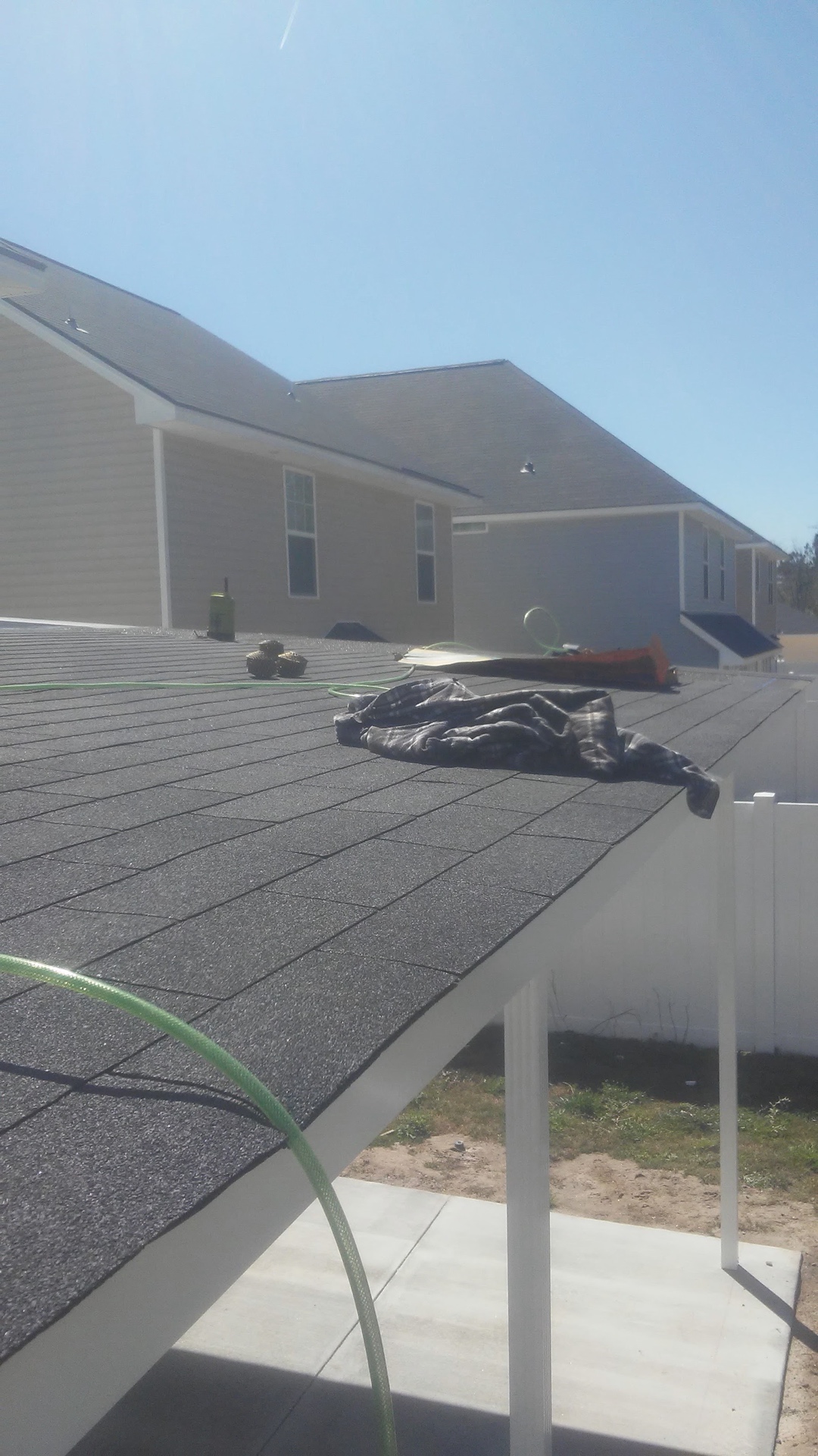 Looking low across the newly shingled dark gray roof, a garden hose and the crew’s tools still resting on it, the neighboring houses beyond