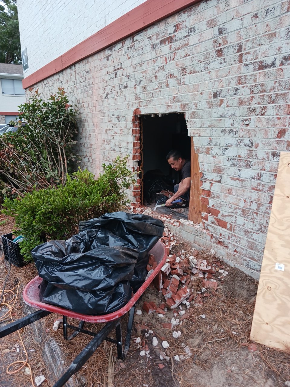 Mid-repair — a worker inside the opening cut through the white-painted brick wall, with bagged debris, loose brick and a sheet of plywood on the ground in front of it