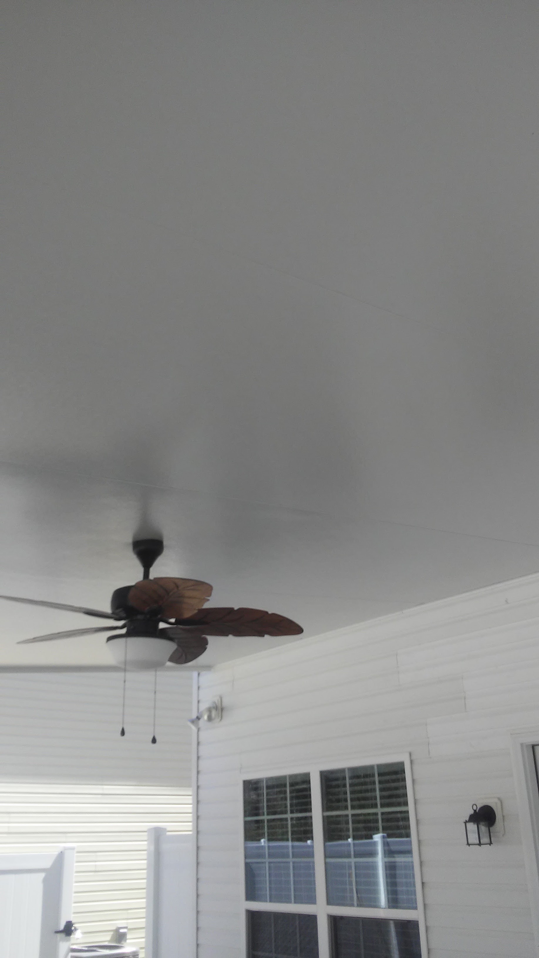 The ceiling fan with dark leaf-shaped blades and a light in the middle, mounted on the white ceiling of the covered patio