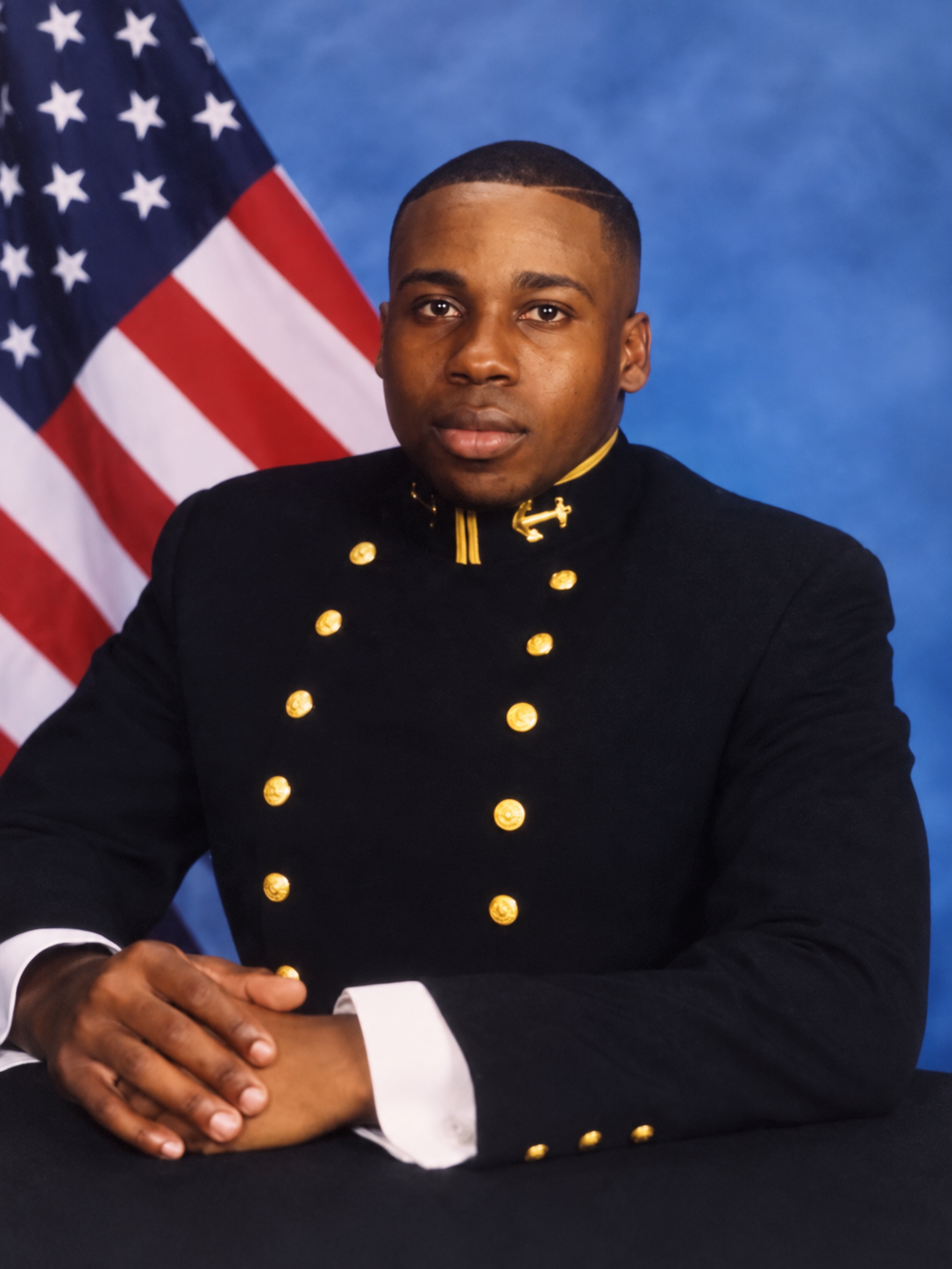 Rod seated in a dark U.S. Navy uniform with gold buttons and a gold anchor insignia on the collar, his hands clasped, the American flag behind him — from his time at the United States Naval Academy in Annapolis, Maryland