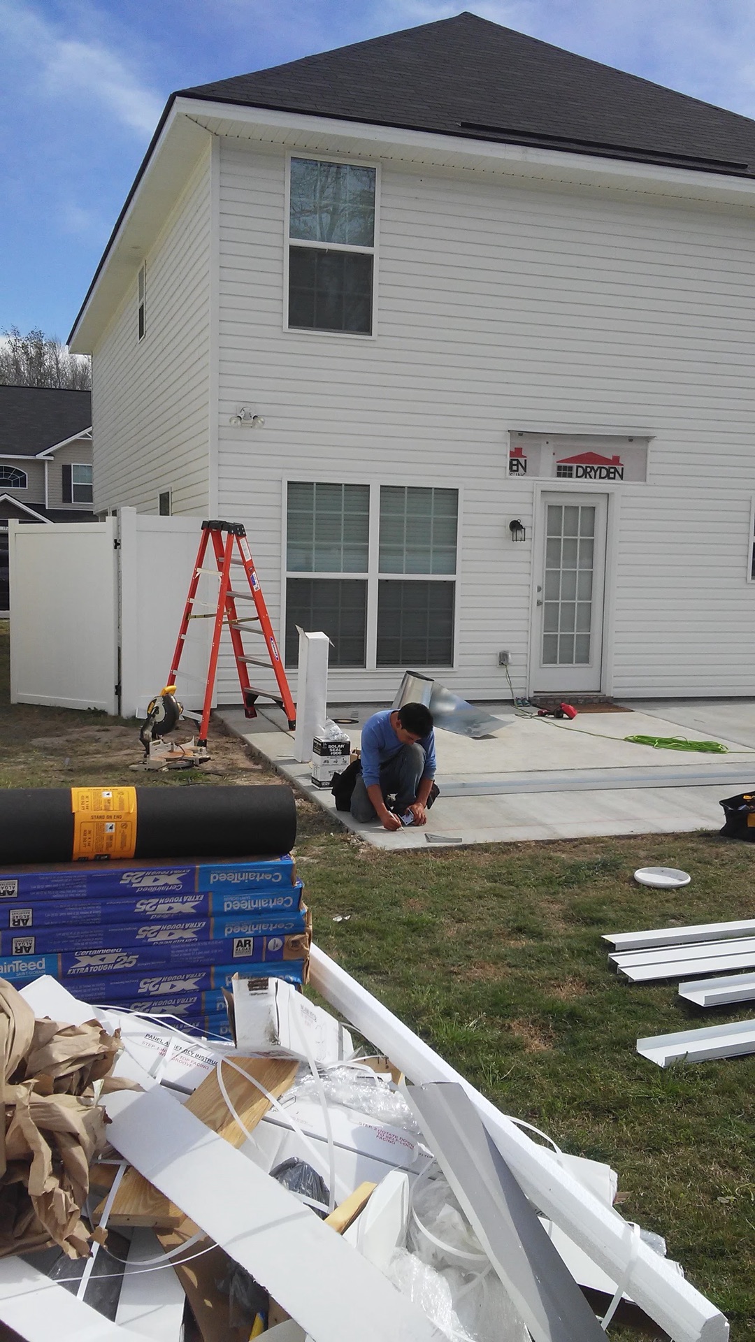 Kneeling at the slab edge to work the metal flashing in against the siding, with the ladder up and insulation, roofing roll and white trim staged on the grass