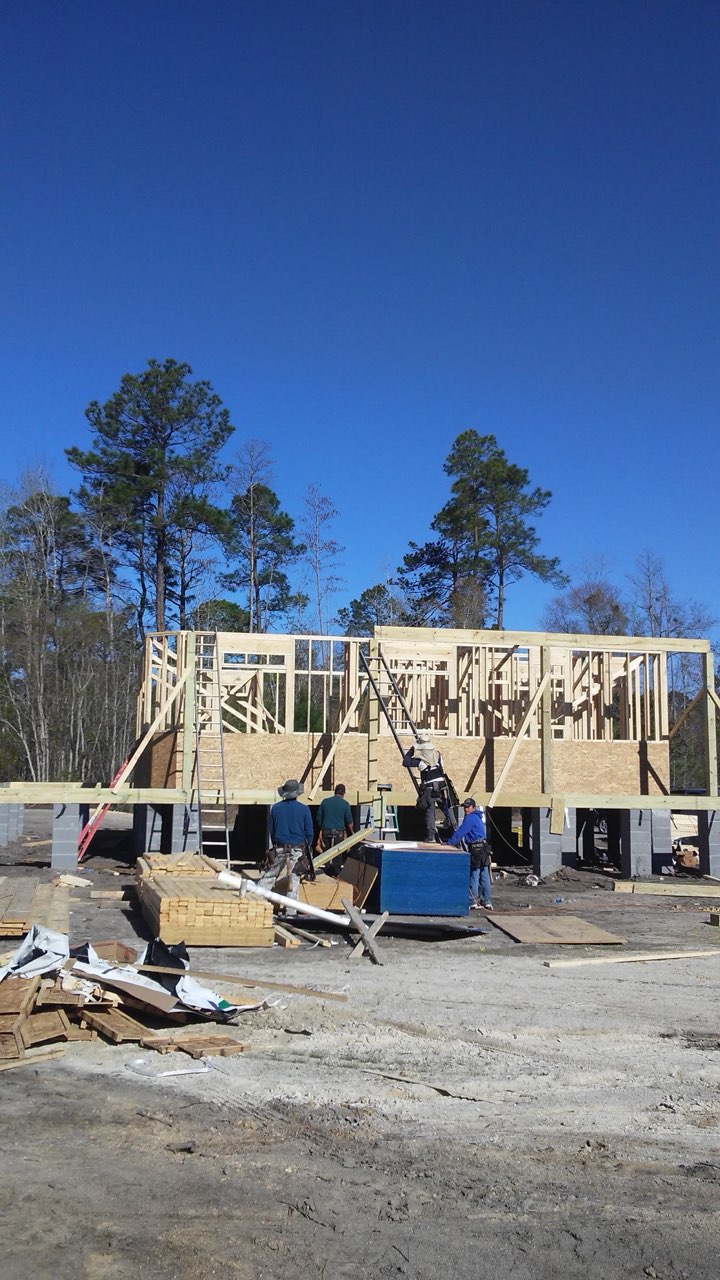 New home framed up on concrete cinder-block foundation piers, lumber staged and pines behind