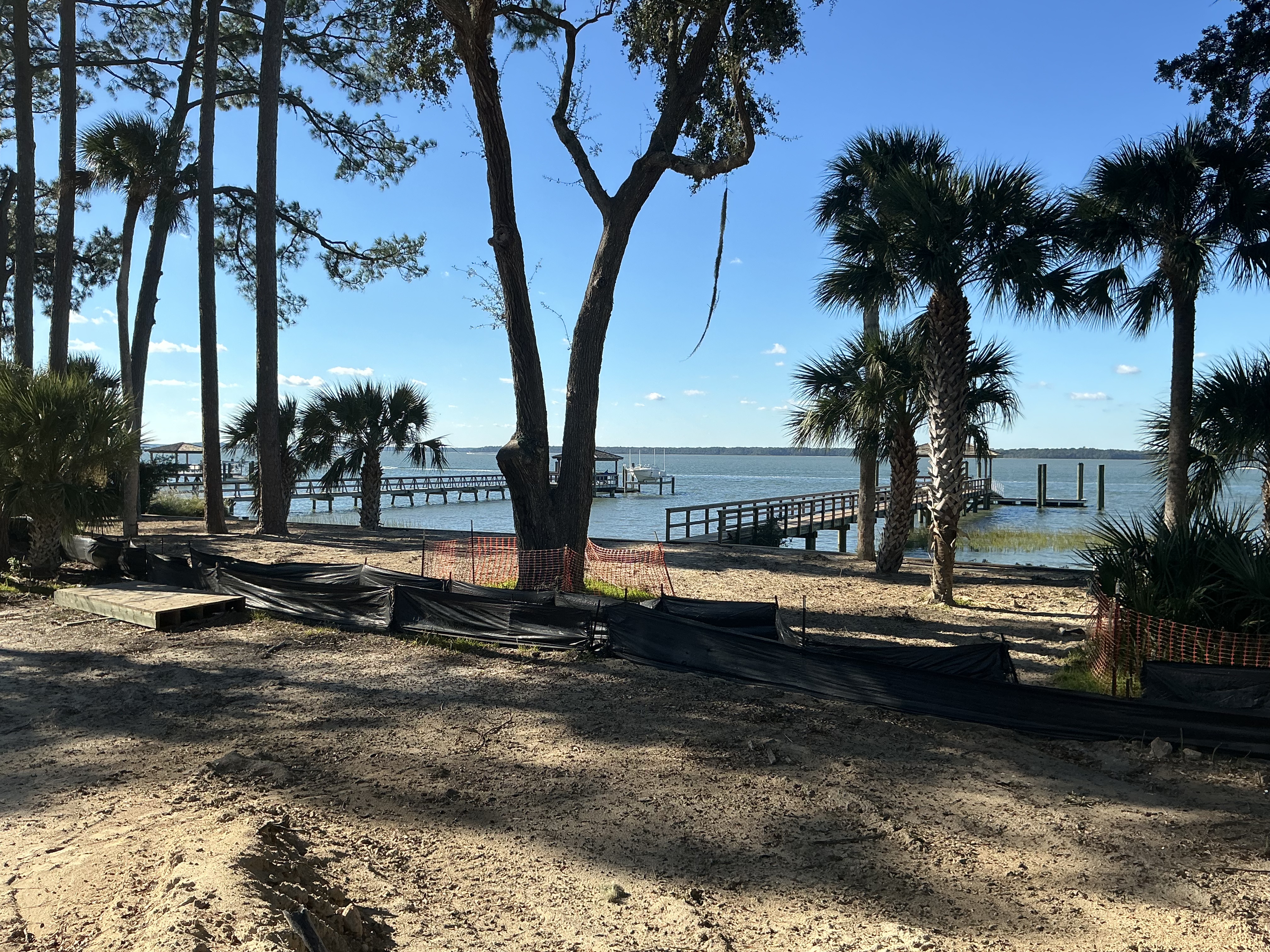 Low Country waterfront lot under work — pines and palms, silt fencing, and docks reaching into the bay