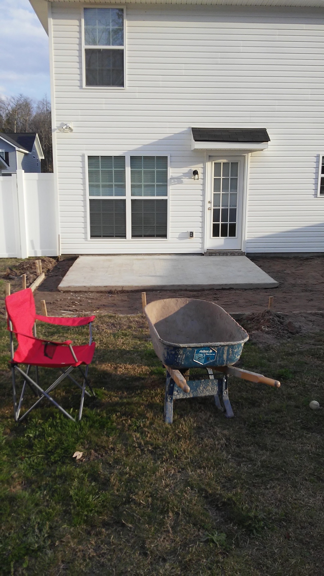 Bare ground behind the white two-story house — a wheelbarrow and a red folding chair on the lawn in front of the small existing pad
