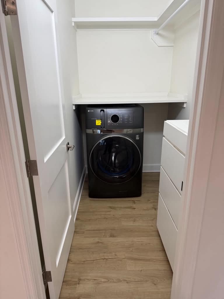 Finished closet — the black front-load washer-dryer unit built in under a white shelf with a hanging rod above and a storage drawer unit beside it, the vinyl plank floor showing across the nook