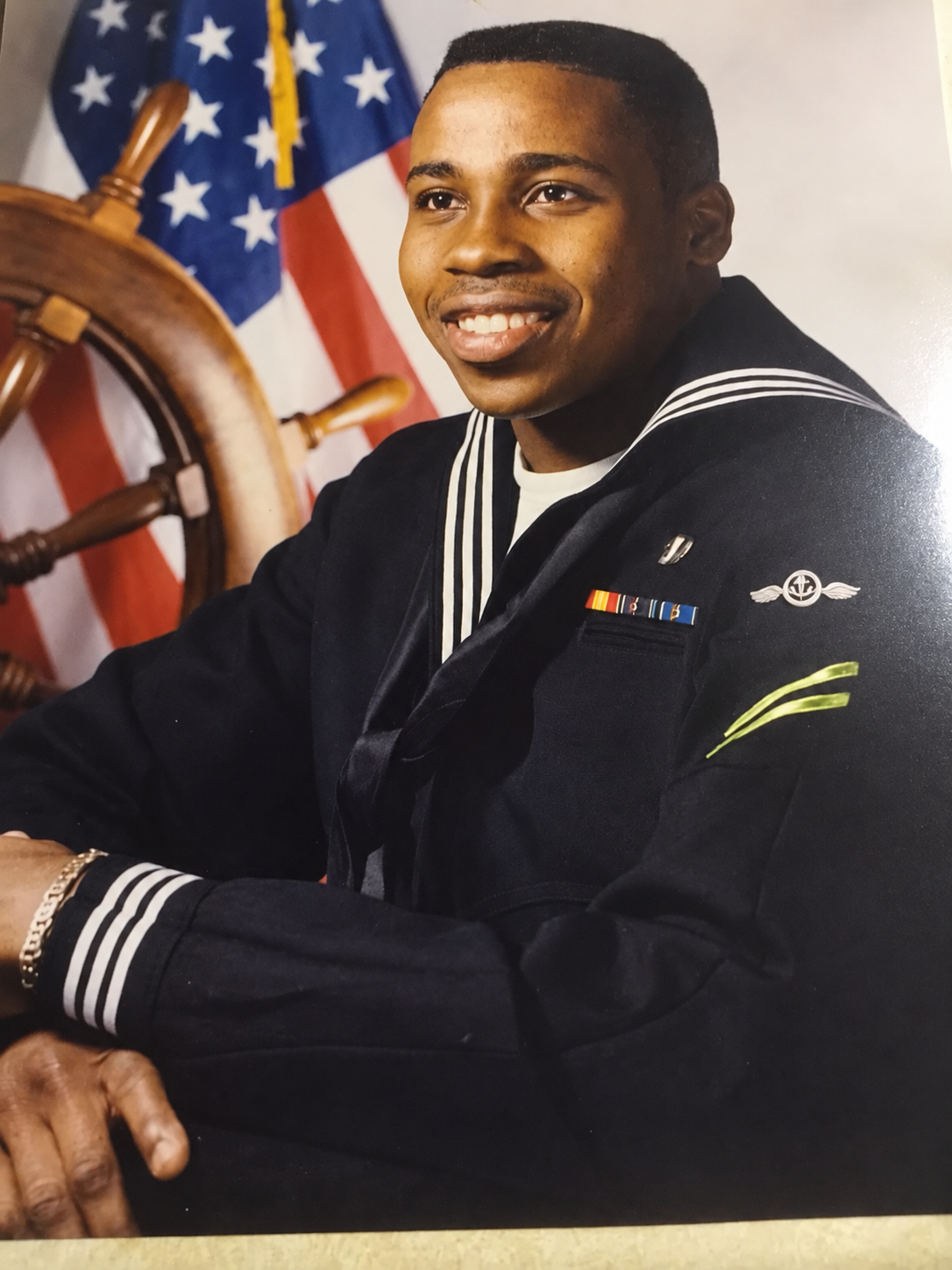 Rod Milledge smiling in a dark blue U.S. Navy dress uniform with ribbons and his silver basic parachute wings and aircrew insignia reading clearly on his chest, posed in front of the American flag and a ship’s wheel — fresh out of jump school